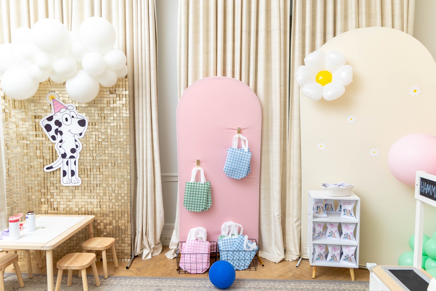 Favor wall with gingham gift bags in pink, blue, and green hung from gold hooks against a pink arched backdrop