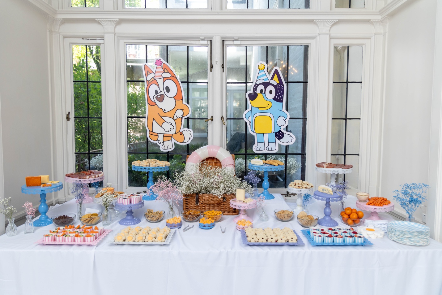 Grazing table with charcuterie, mini sandwiches, fruit, Bluey sugar cookies, and a tiered cake with pastel balloon cascade