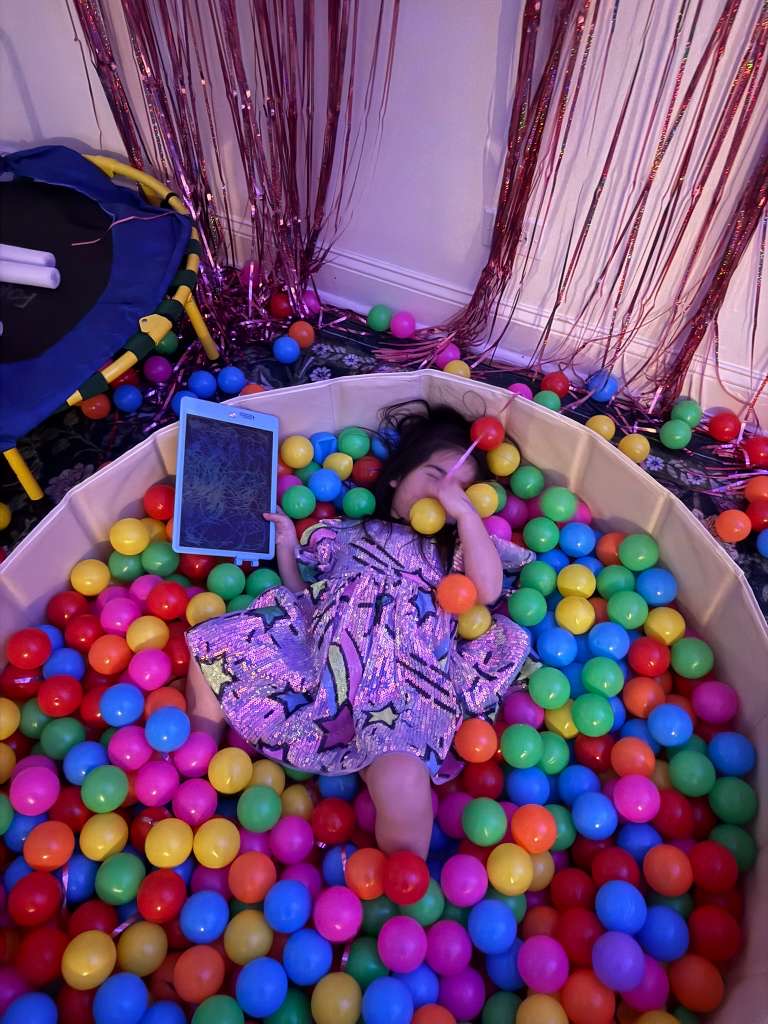 Roxy lying in the ball pit with a tablet, surrounded by colorful balls and foil fringe curtains