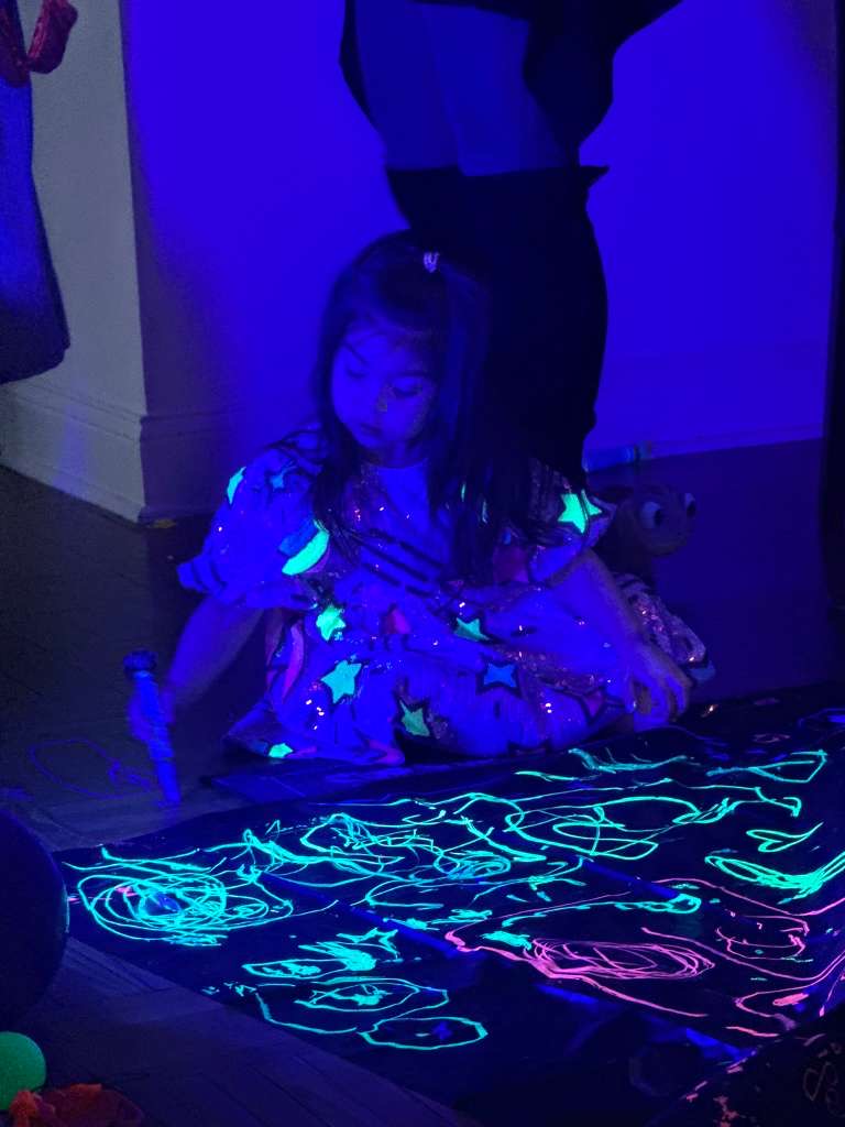 A child sitting under blacklight with glowing neon art covering the tarp in front of her