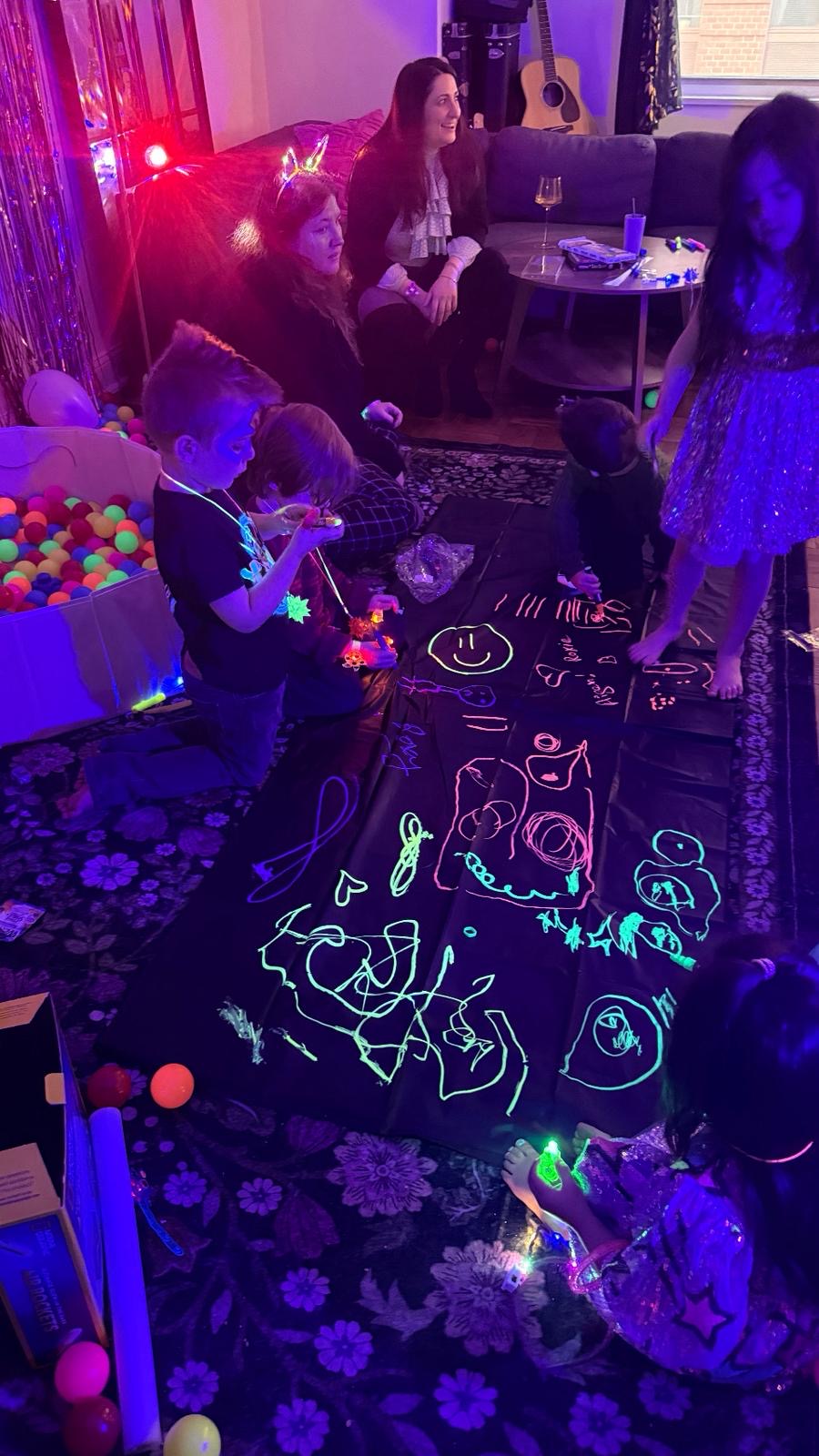 Kids and adults gathered around the glow art tarp — neon drawings glowing under blacklight, ball pit visible in the background