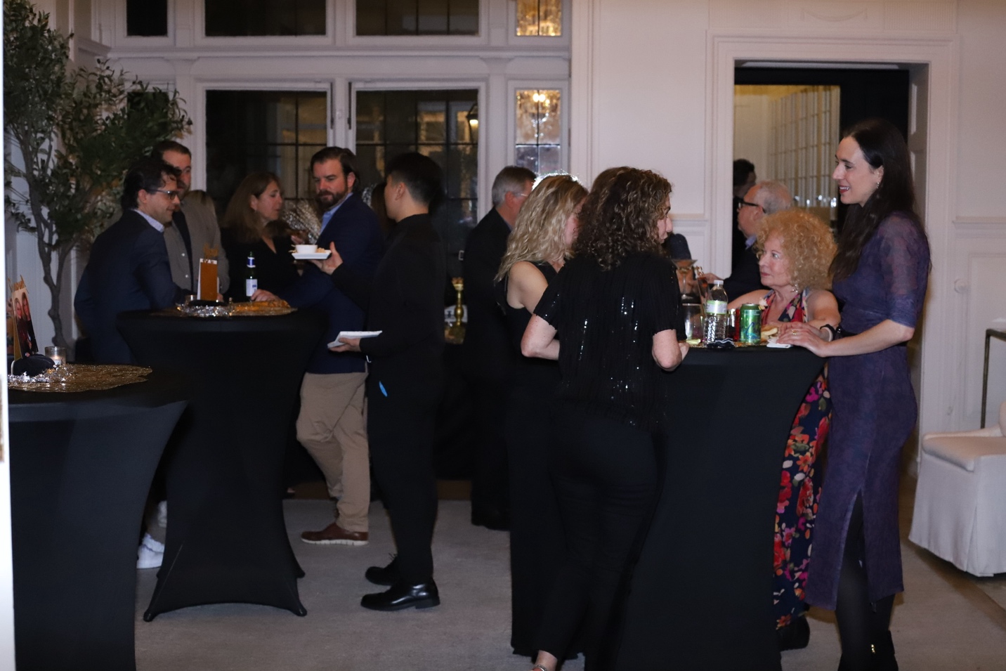 Guests mingling at cocktail tables during the party — black tablecloths, olive trees, and warm lighting
