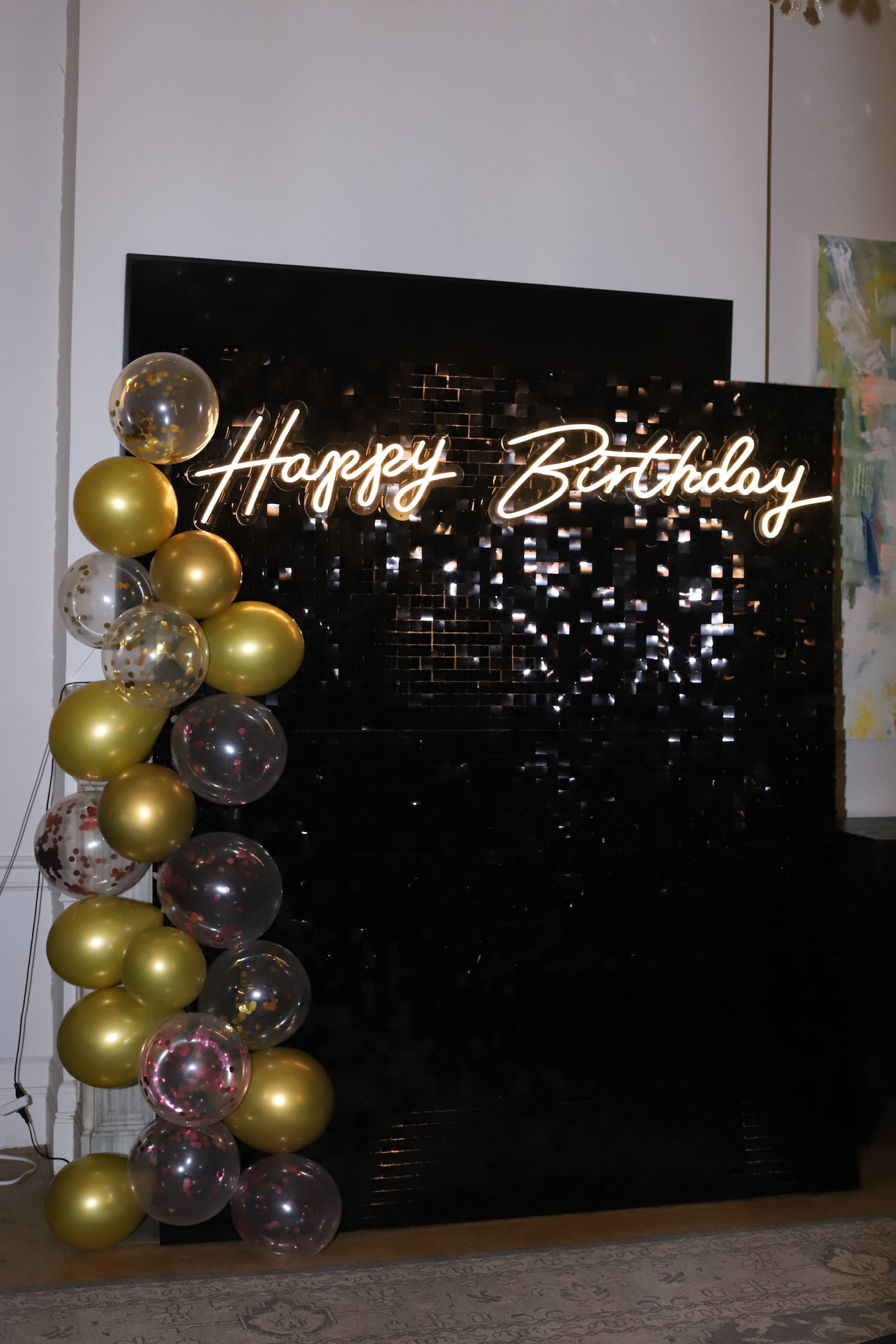 Black sequin backdrop with warm white Happy Birthday neon sign and gold and confetti balloon column
