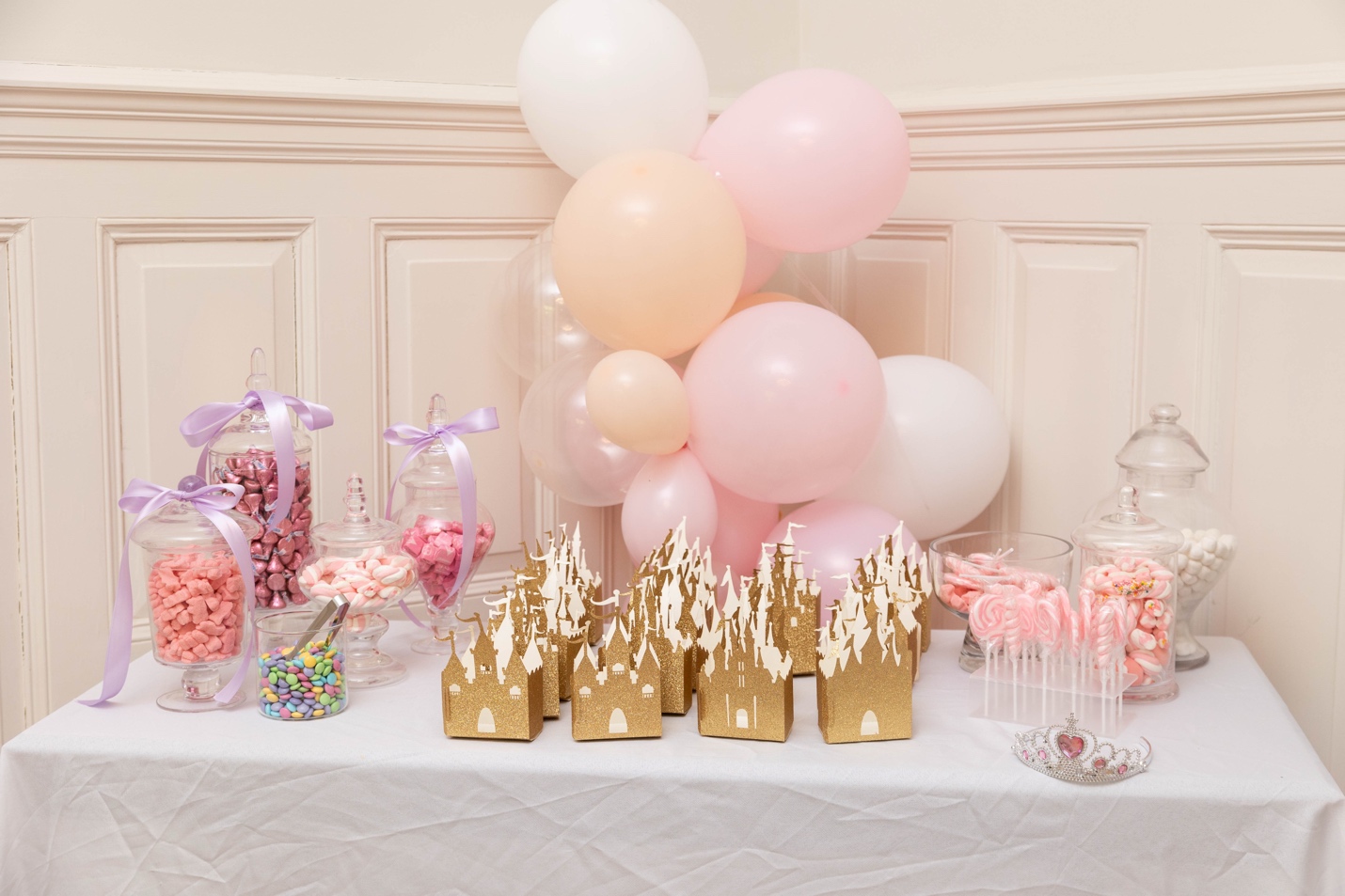 Candy bar station with glass apothecary jars, gold castle favor boxes, pastel balloon garland, and a tiara
