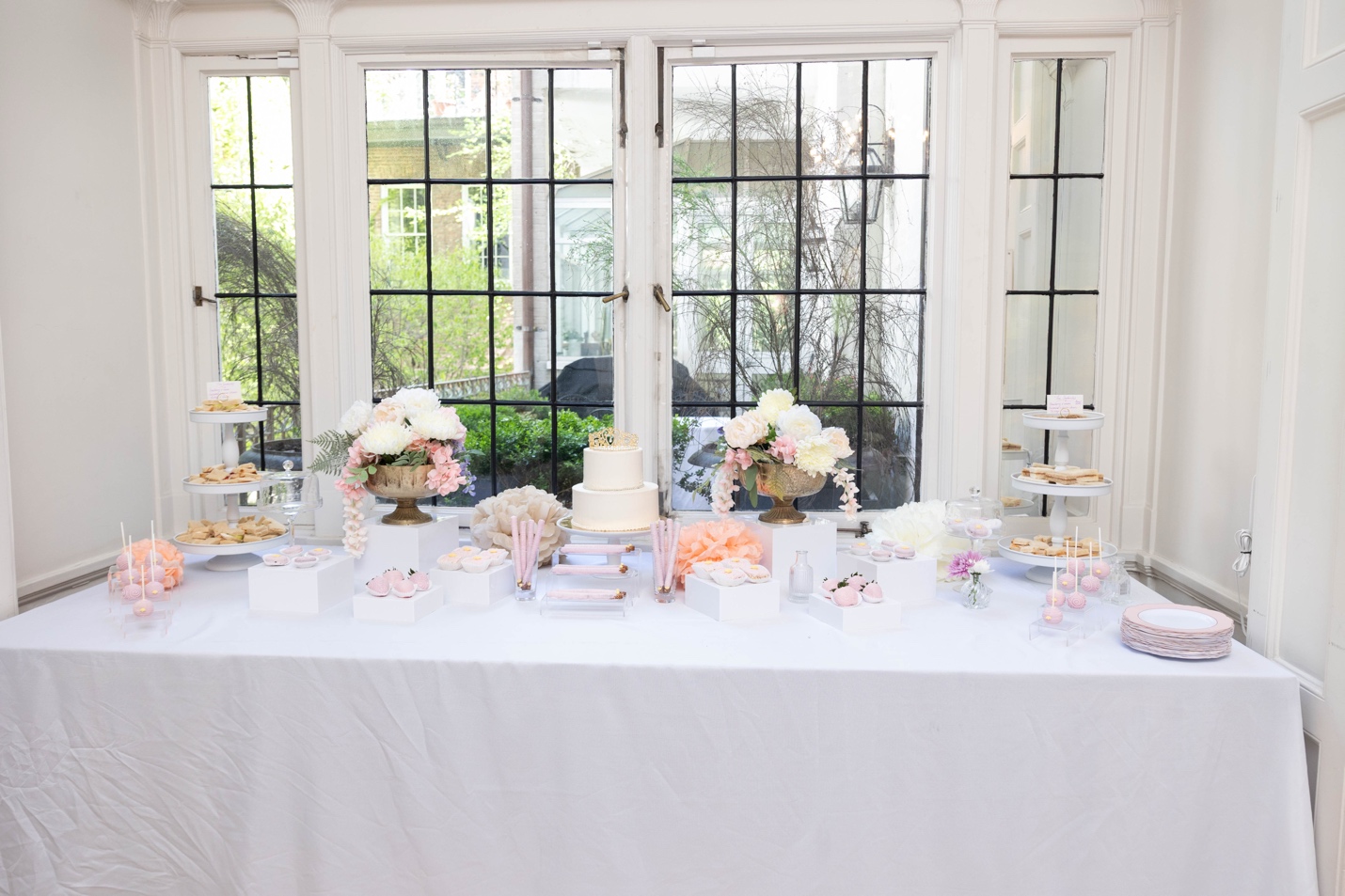 Dessert table against floor-to-ceiling windows — two-tier cake, tiered trays, cake pops, floral arrangements in gold urns
