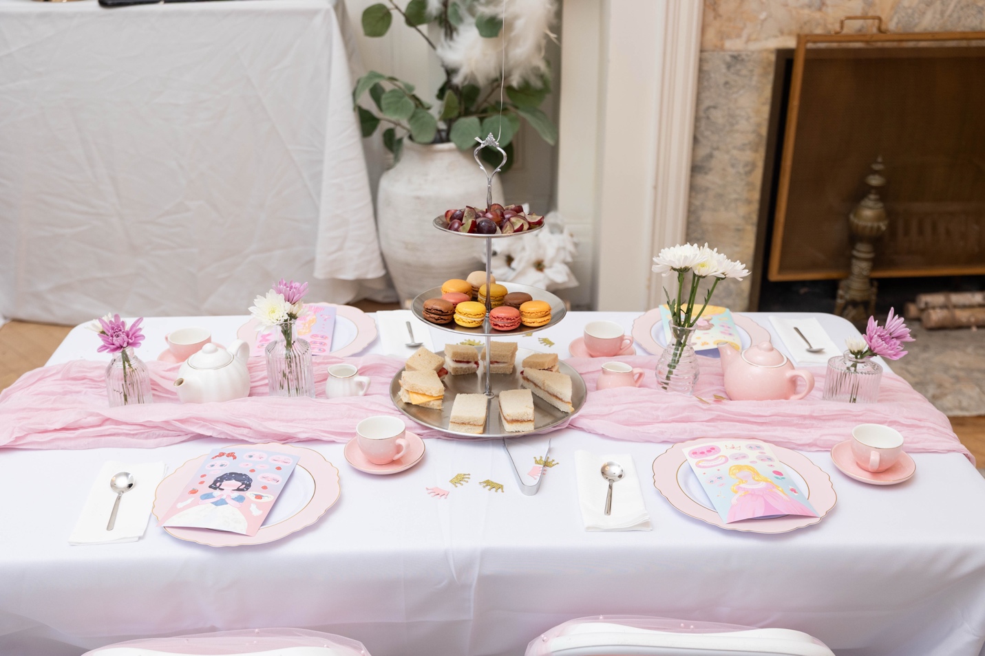 Close-up of a single table setting — porcelain teacups, tiered stand with macarons and finger sandwiches, bud vases with fresh flowers